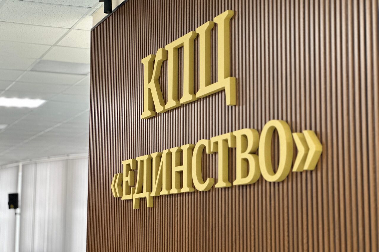 Арендовать КПЦ Единство. Стена с вертикальными деревянными панелями, украшена объемными золотыми буквами с текстом "КПЦ ЕДИНСТВО". На заднем плане видна часть потолка с плафоном и колонна.
