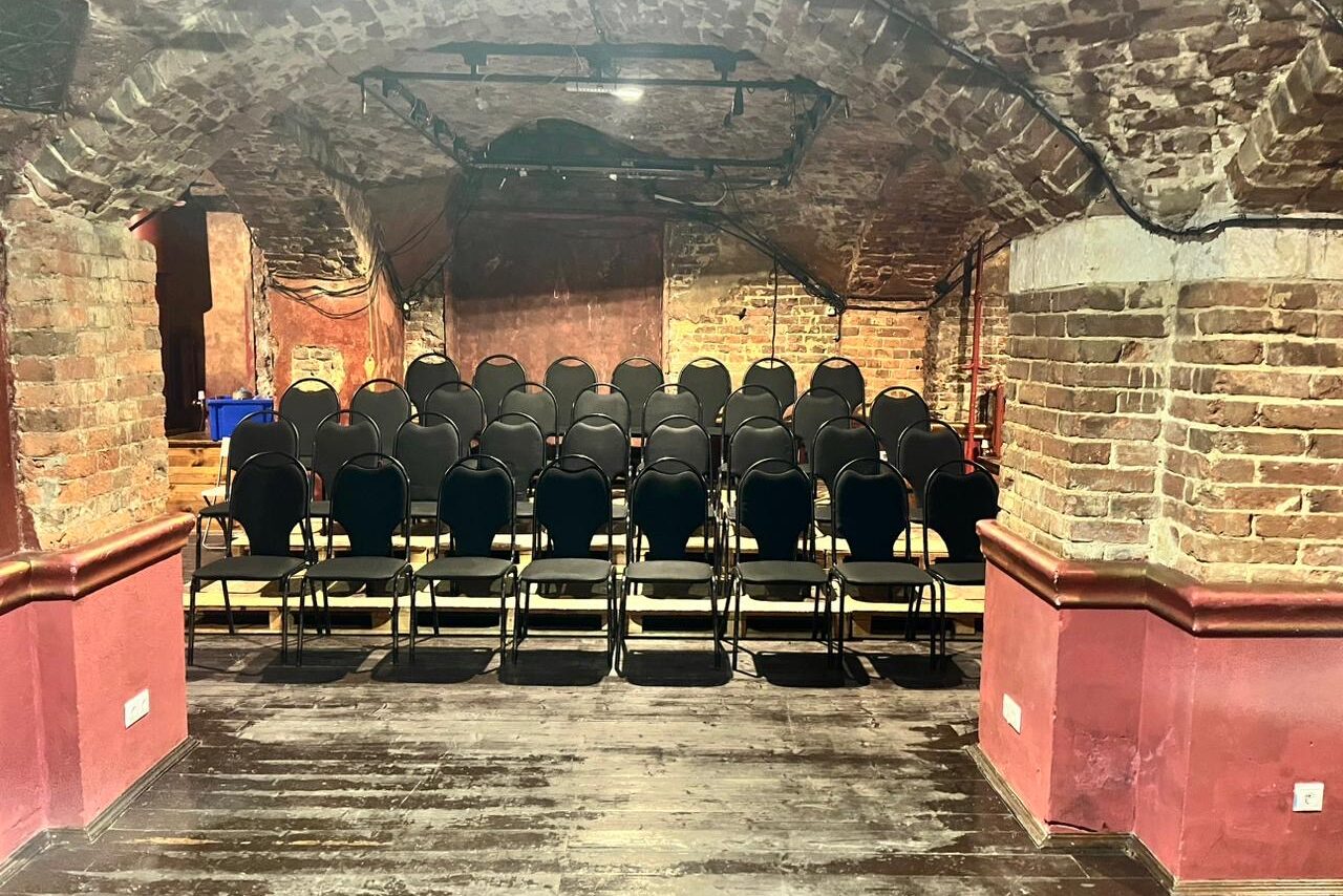 Арендовать Культурный центр «Хитровка». "Арт-зал". Старинное подвальное пространство с кирпичными арками и каменными стенами, темный деревянный пол, черные стулья в ряды, мягкое освещение сверху, бордовые панели на стенах, черные трековые светильники.