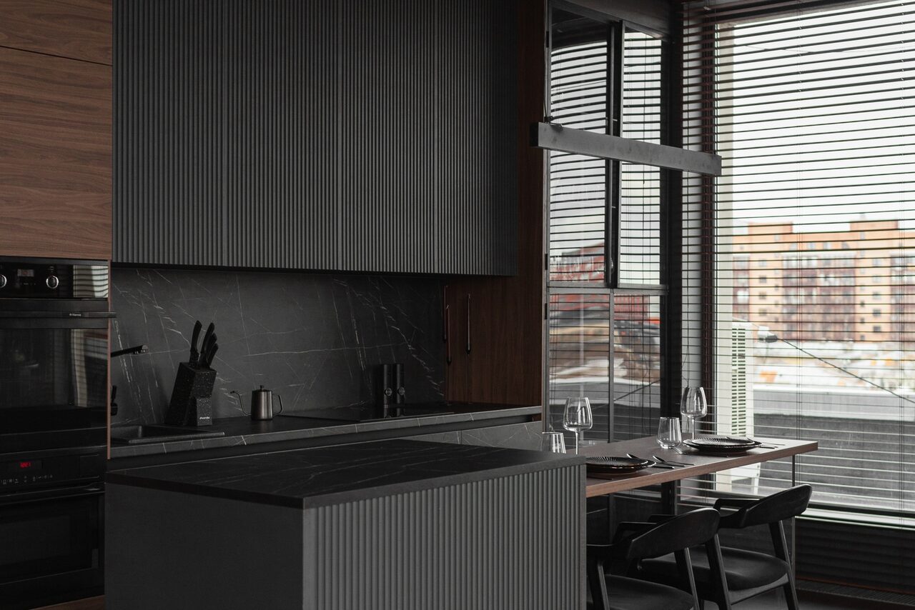 Арендовать Зал Black. Фотостудия Мимика. Современная кухня с серыми и черными элементами, барной стойкой и столом у окна. На столе три стеклянных бокала и приборы. Вертикальные жалюзи на окнах. Тёмные шкафы с деревянными вставками. Слева у стены плита и раковина.