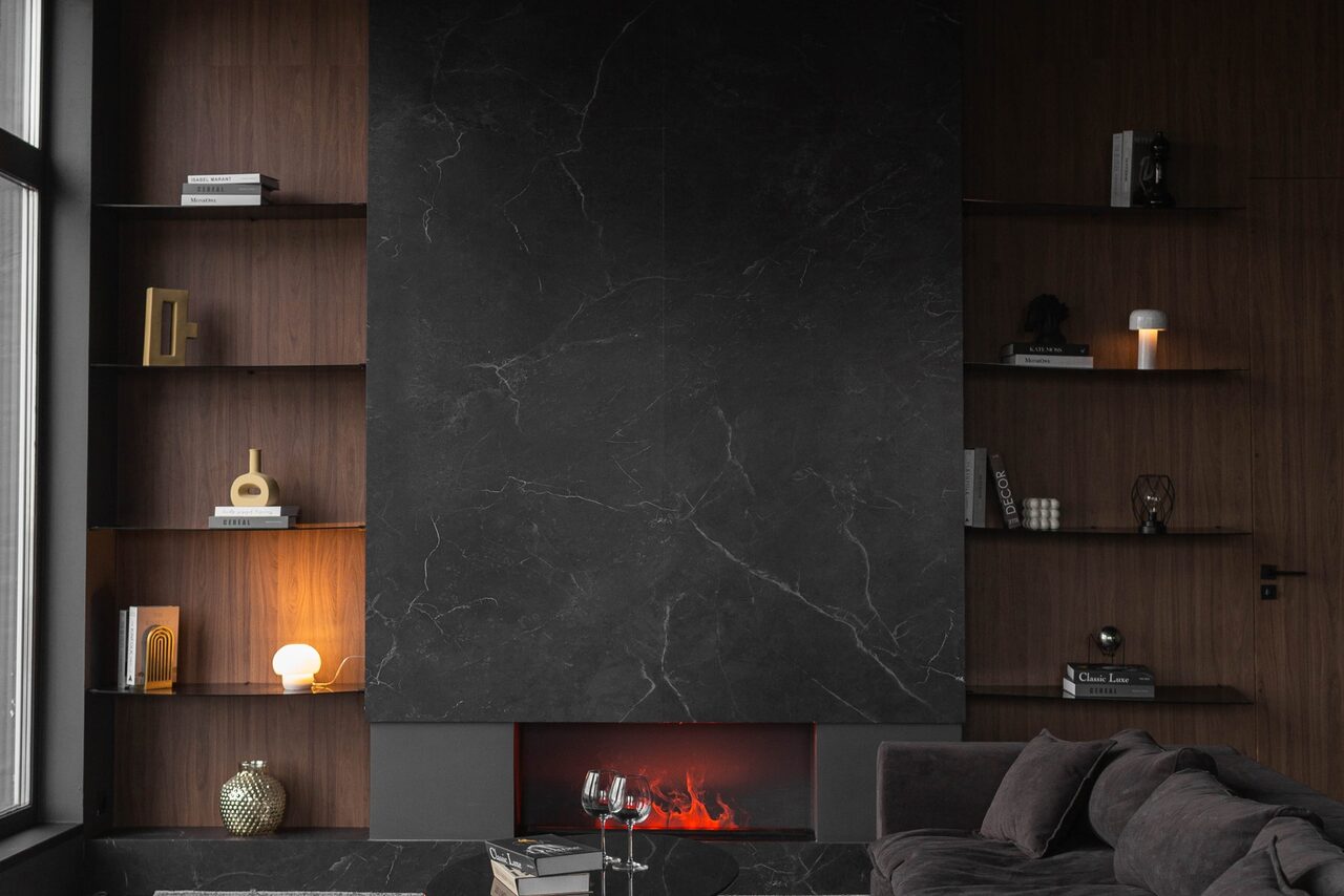 Арендовать Зал Black. Фотостудия Мимика. Гостиная с камином. По бокам камина расположены полки с книгами и декоративными элементами. На полу стоит стеклянный столик с бокалами. Справа тёмный диван. Стены облицованы тёмным деревом и чёрным мрамором. Освещение создают настольные лампы и окно слева.
