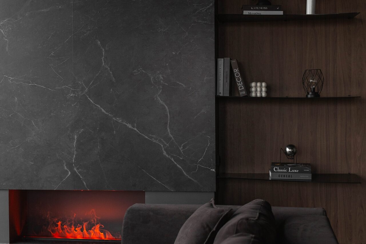 Арендовать Зал Black. Фотостудия Мимика. Современная гостиная с черным мраморным камином, создающим эффект пламени. Справа деревянные полки с книгами и декором. Спереди темный диван с подушками. Темные, нейтральные оттенки и минималистичный стиль.