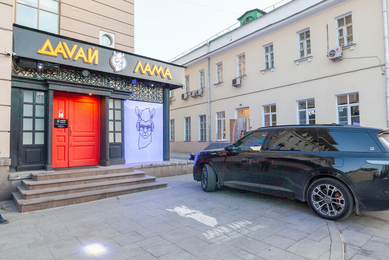 Арендовать Давай Лама. Вход в здание с красной двойной дверью, стеклянными вставками и черной отделкой. Вывеска на русском языке. Ступени ведут к двери. Черный автомобиль припаркован справа. На стене граффити с изображением ламы.