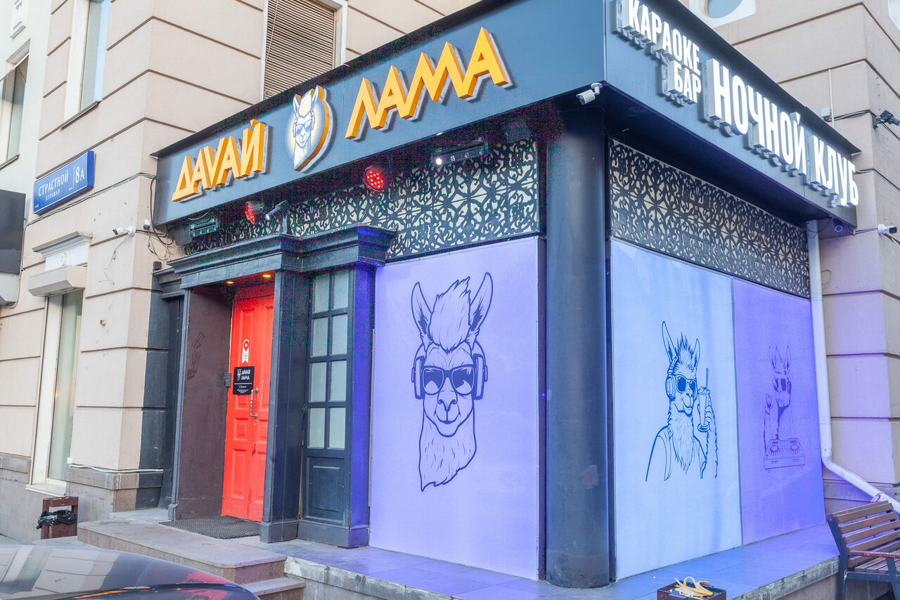Арендовать Давай Лама. Фасад здания с вывеской над входом; красная дверь, графические изображения ламы в наушниках на фиолетовых панелях; надписи на русском языке: "Да-Вай Лама", "Караоке бар Ночной клуб"; архитектура двухуровневая; декоративная решетка.