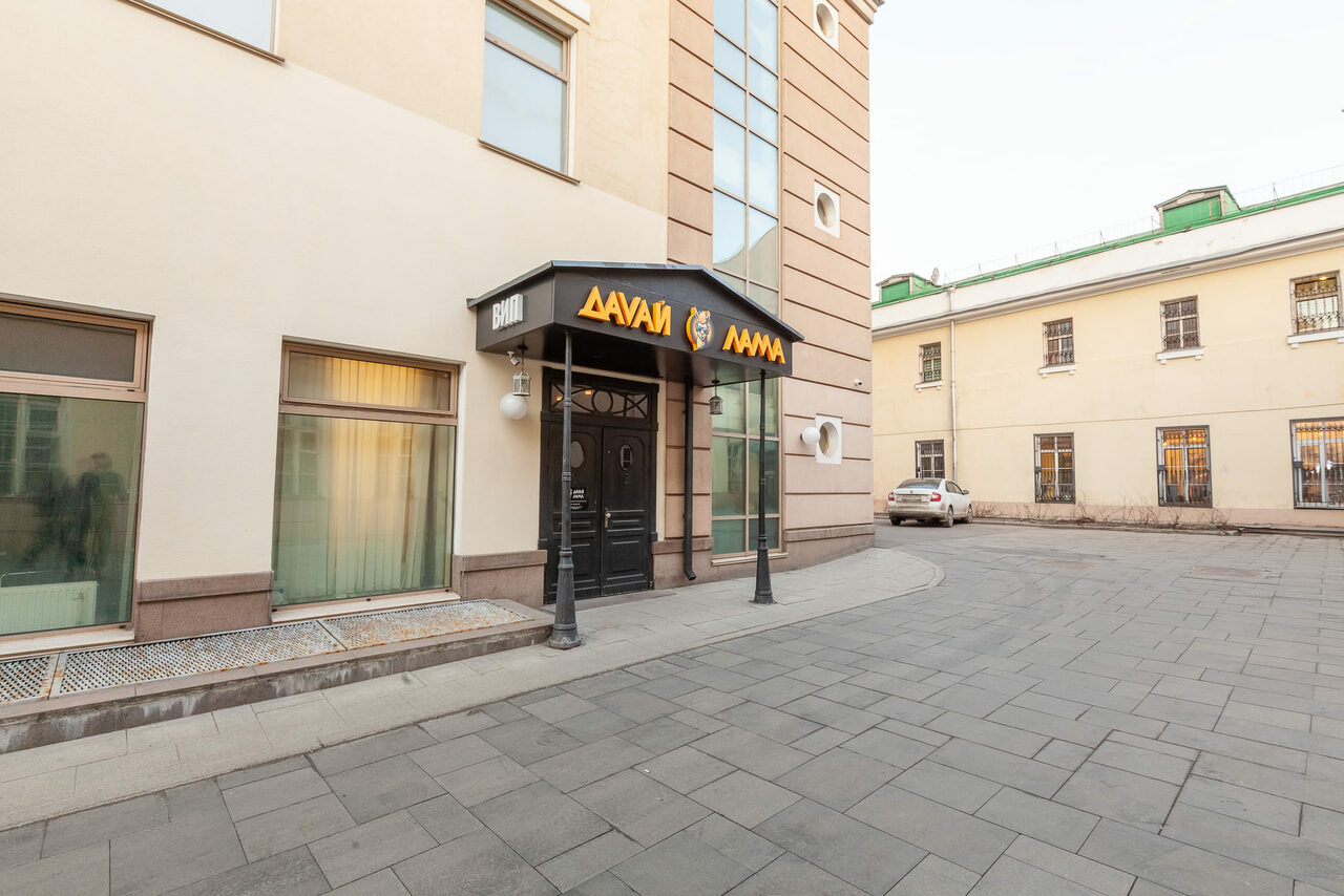 Арендовать Давай Лама. Многоэтажное здание с бежевым фасадом и большими окнами. Перед входом черный козырек с желтой вывеской на неизвестном языке. Входная дверь темного цвета. Асфальтированная территория с парковкой, виден автомобиль.
