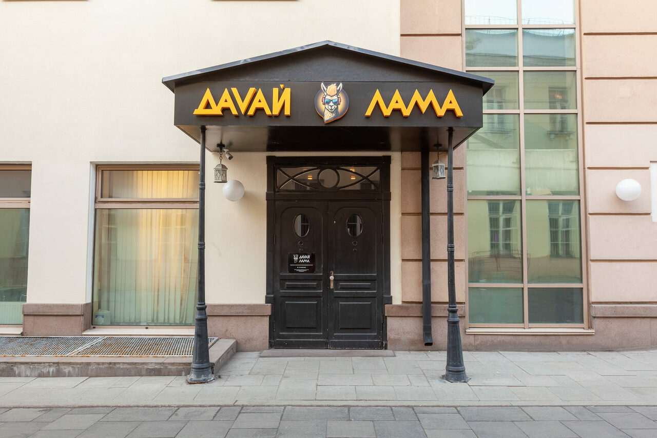 Арендовать Давай Лама. Вход в здание с черными двустворчатыми дверями под черным навесом. Светлый фасад с большими окнами по бокам. Черные фонари подвешены под навесом. На крыше навеса желтая вывеска с текстом на кириллице.