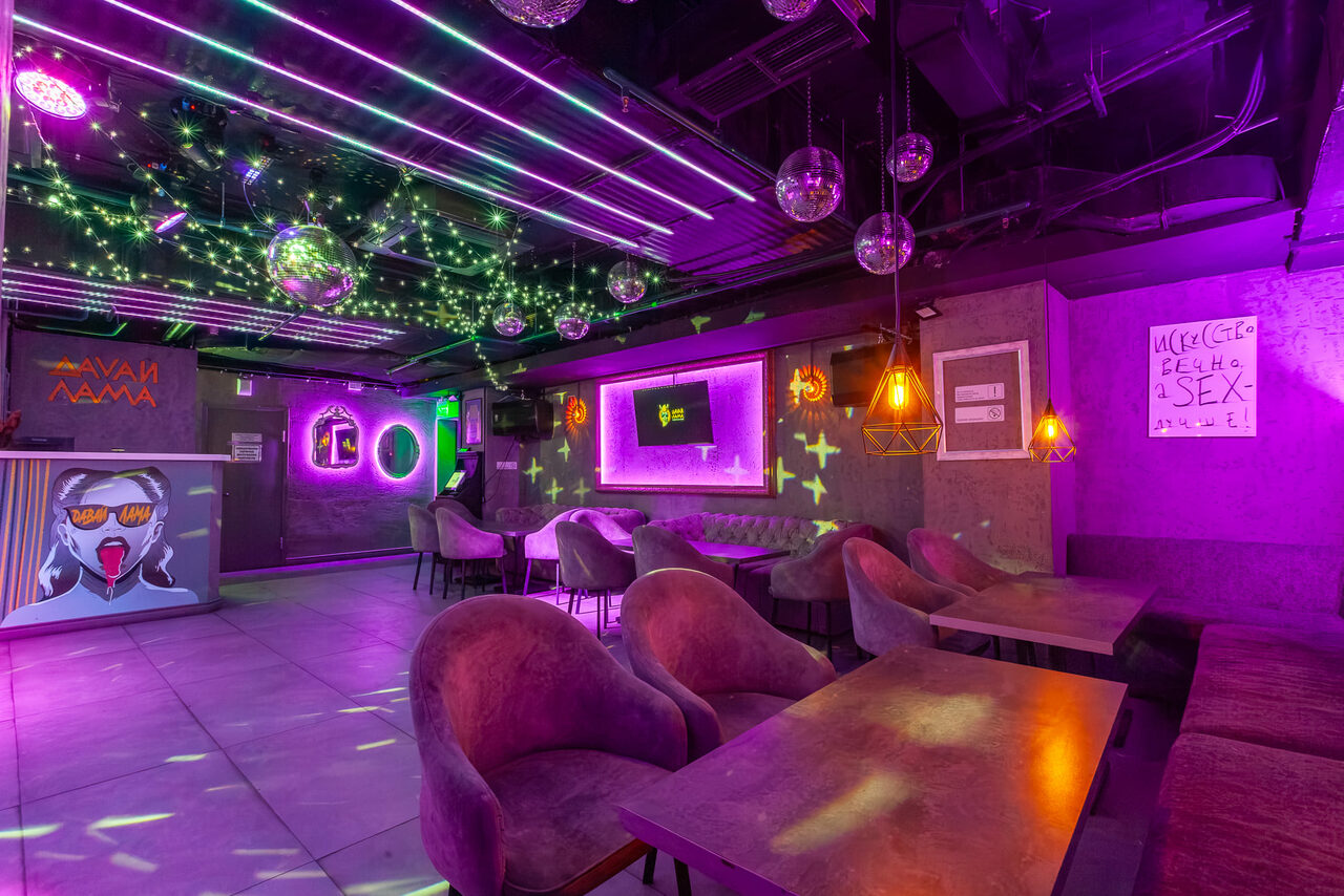 Арендовать Давай Лама. Loft space with neon and purple lighting. Features include round tables, velvet chairs, and wall art. Multiple pendant lights and disco balls hang from the ceiling. Walls are concrete, some with graffiti. Ceiling has illuminated strips and star-like patterns.