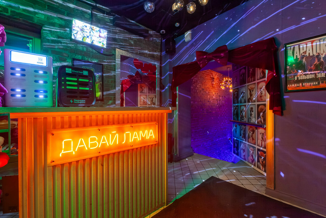 Арендовать Давай Лама. Караоке-бар с кирпичной стеной, стойкой с неоновой надписью, плиточным полом и цветными световыми эффектами. На стенах висят плакаты, рядом с барной стойкой — оборудование и экраны. Потолок украшен светильниками.