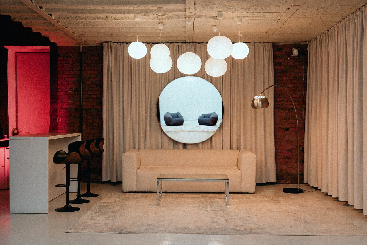 Арендовать Ö Art Space(Арт Спейс). Современное арт-лофт пространство. Лофт с кирпичными стенами и белым потолком. В центре диван, стеклянный стол, круглый настенный элемент с изображением подушек. Справа торшер, слева барная стойка и четыре стула. Потолочные светильники-шары, занавесы светлого цвета.