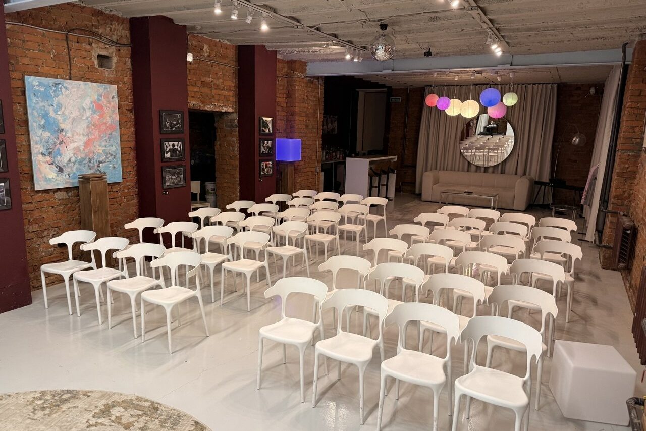 Арендовать Ö Art Space(Арт Спейс). Современное арт-лофт пространство. Просторное помещение с кирпичными стенами, оборудованное белыми стульями, расположенными в ряд. На стене висит яркая картина, рядом с ней множество фотографий. Зал украшен разноцветными подвесными шарами, на фоне зеркало и диван. Потолок со светильниками.