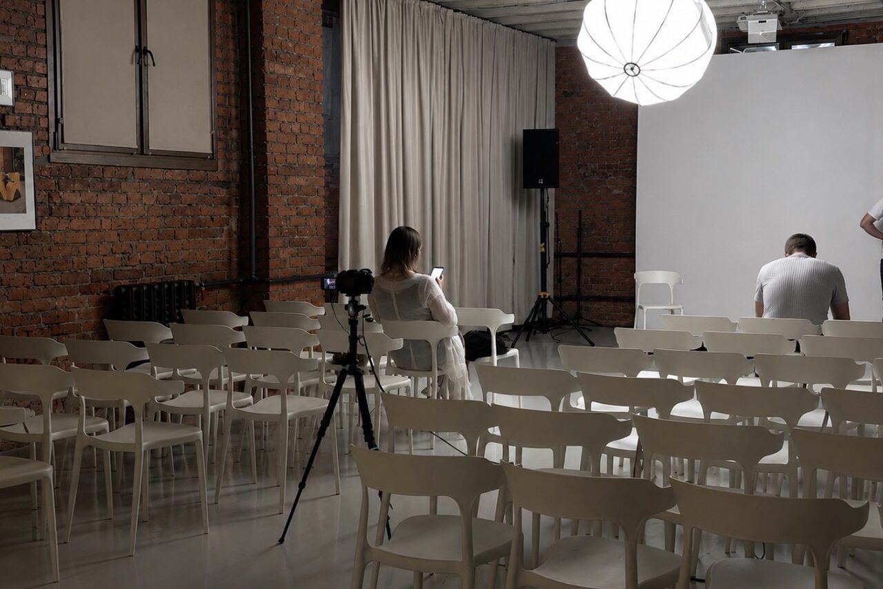 Арендовать Ö Art Space(Арт Спейс). Современное арт-лофт пространство. Лофт-пространство с кирпичными стенами, белыми стульями, расположенными рядами. Освещение от крупного подвесного светильника. На стене картинная рамка. Белая занавеска, светлая напольная поверхность. На штативе установлен фотоаппарат.
