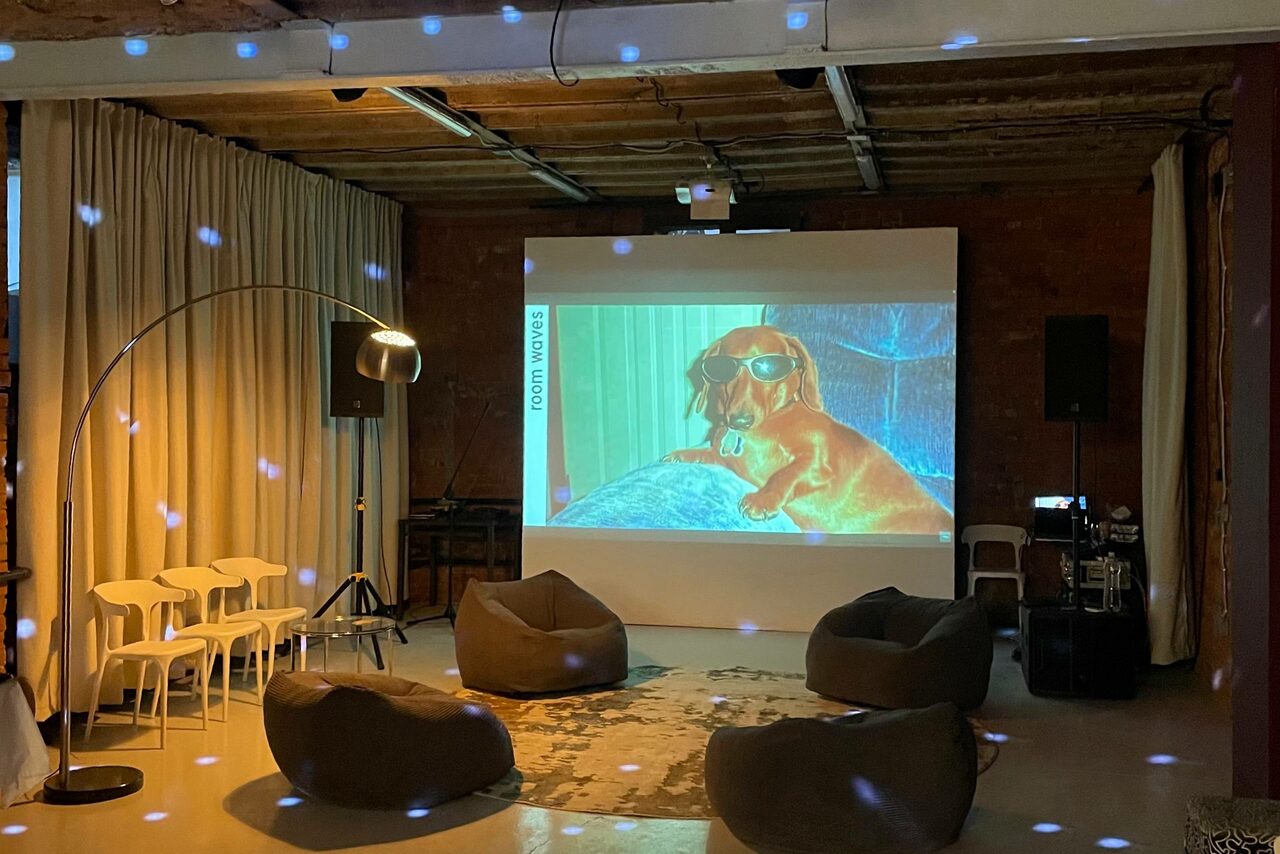 Арендовать Ö Art Space(Арт Спейс). Современное арт-лофт пространство. Лофт-пространство с кирпичными стенами и открытым потолком. В центре проектор с изображением собаки в очках. Мягкие кресла, белые стулья, торшер, светодиодное освещение. Шторы и ковёр светлых тонов. Звуковое оборудование, стол, провода.