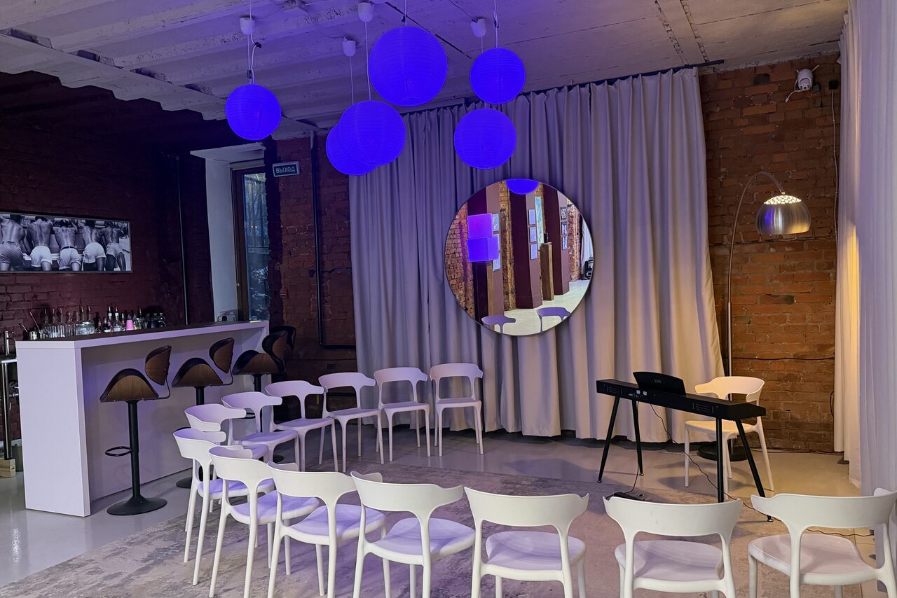 Арендовать Ö Art Space(Арт Спейс). Современное арт-лофт пространство. Пространство с кирпичными стенами, барной стойкой и рядом высоких стульев. Полукруг из белых стульев перед клавишным инструментом. На стене круглое зеркало и изображение. Подвесные круглые светильники фиолетового цвета, занавески.