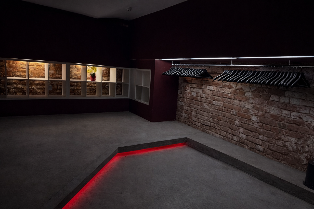 Арендовать Ö Art Space(Арт Спейс). Современное арт-лофт пространство. Подвал с кирпичными стенами и полками с подсветкой. На одной стене висят пустые вешалки на рейле. Пол из серого бетона с выступом, с красной LED-подсветкой. Потолок темный. Полки с цветочной декорацией внутри.