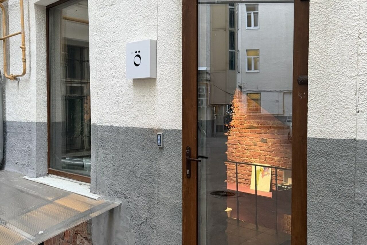 Арендовать Ö Art Space(Арт Спейс). Современное арт-лофт пространство. Вход в здание с дверью из стекла и дерева. Справа кирпичная стена внутри. На стене белый ящик с чёрным логотипом. Окно с левой стороны. Стены снаружи окрашены в серый и белый. Газовая труба слева.