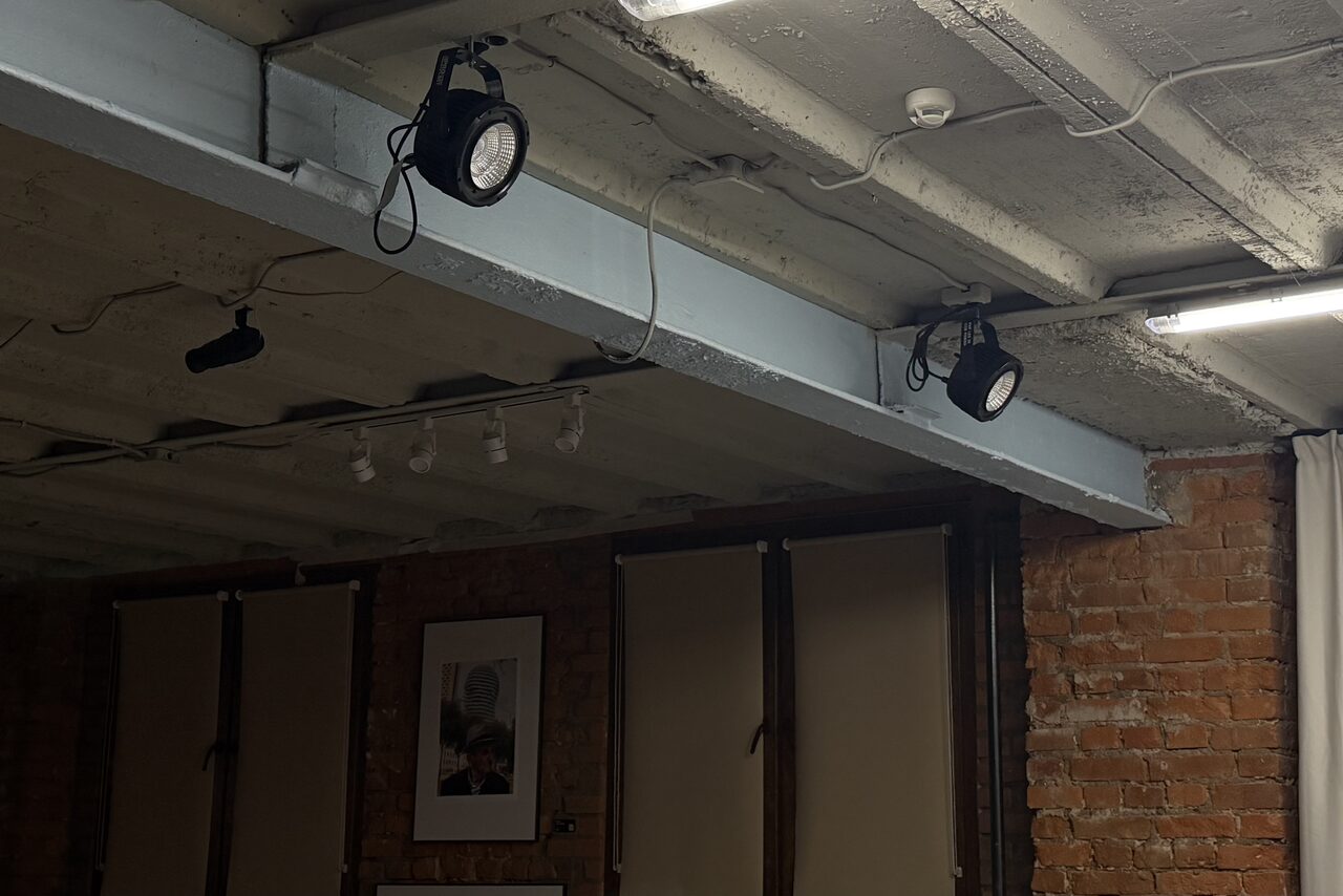 Арендовать Ö Art Space(Арт Спейс). Современное арт-лофт пространство. Лофт с кирпичными стенами и бетонным потолком. Освещение представлено потолочными светильниками и прожекторами. На стене висят картины. Пространство затемнено, свет нейтральный. Картину окружают отсутствующие окна с шторами.
