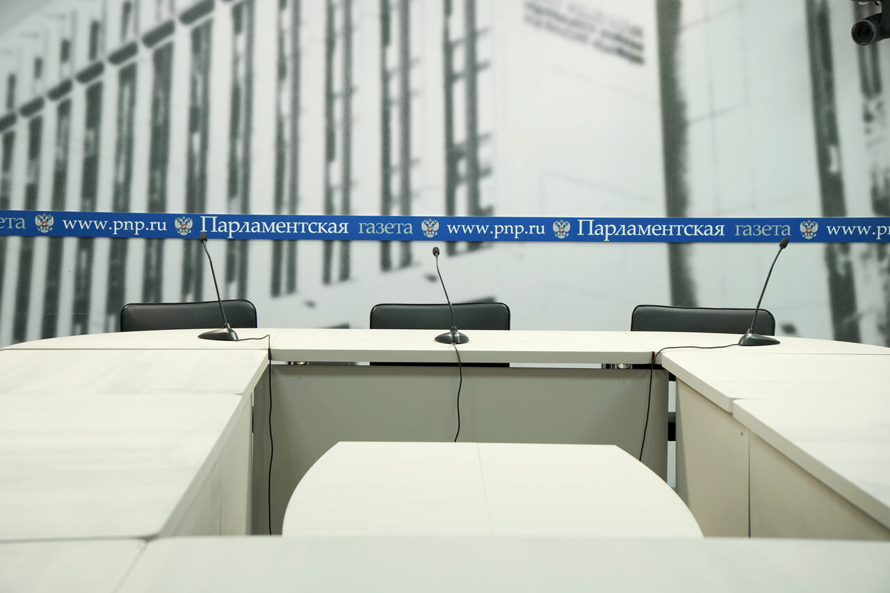 Арендовать Медиацентр Парламентской газеты. Конференц-зал с белыми столами, микрофонами на стойках, черными офисными стульями. На заднем плане серый графический фон с вертикальными линиями. На столах синяя полоса с текстом: "Парламентская газета www.pnp.ru".