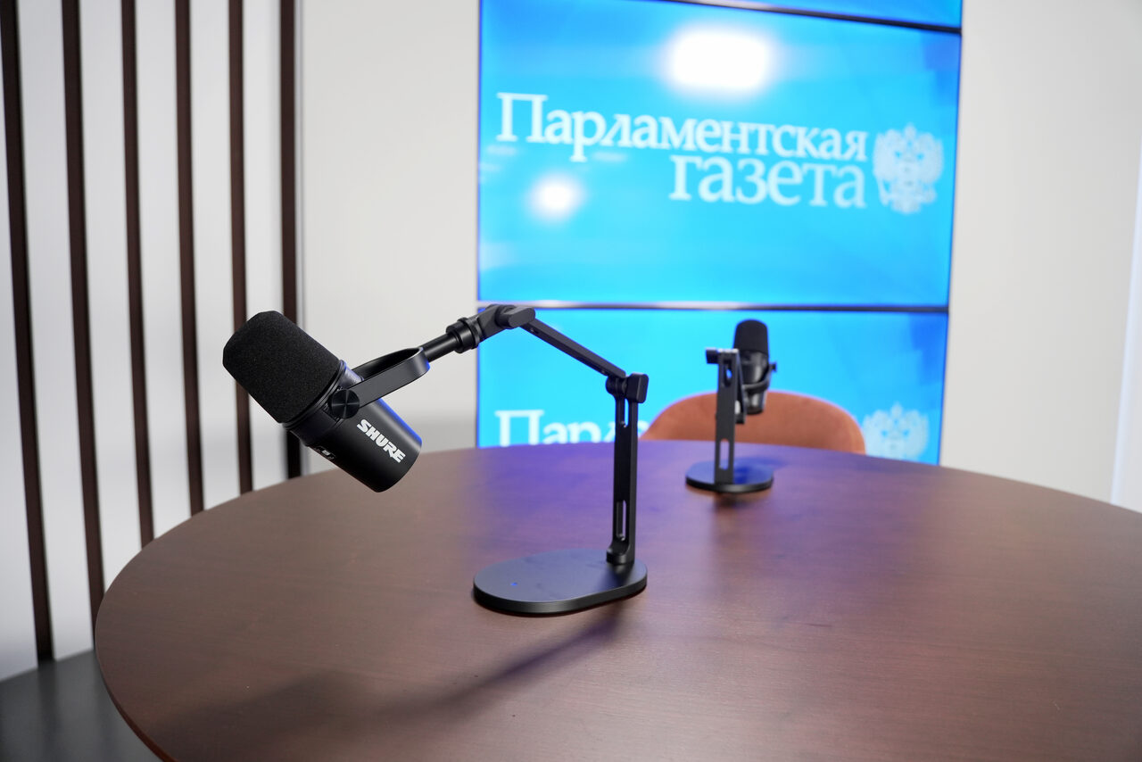 Арендовать Медиацентр Парламентской газеты. Студия для записи подкастов с круглым деревянным столом, двумя микрофонами на стойках и креслом. На заднем плане - синий экран с текстом "Парламентская газета". Стена с вертикальными деревянными панелями.