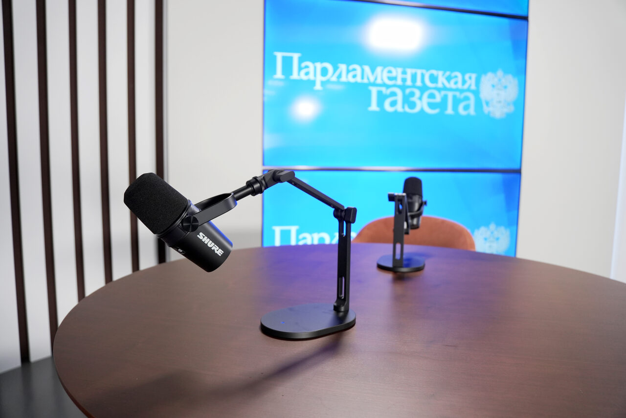 Арендовать Медиацентр Парламентской газеты. Круглый стол с двумя микрофонами на стойках, один стул. Фон с несколькими экранами, на которых видна надпись "Парламентская газета". Вертикальные элементы, похожие на панели или рейки, сбоку. Цвета: коричневый стол, чёрные микрофоны, синий экран.