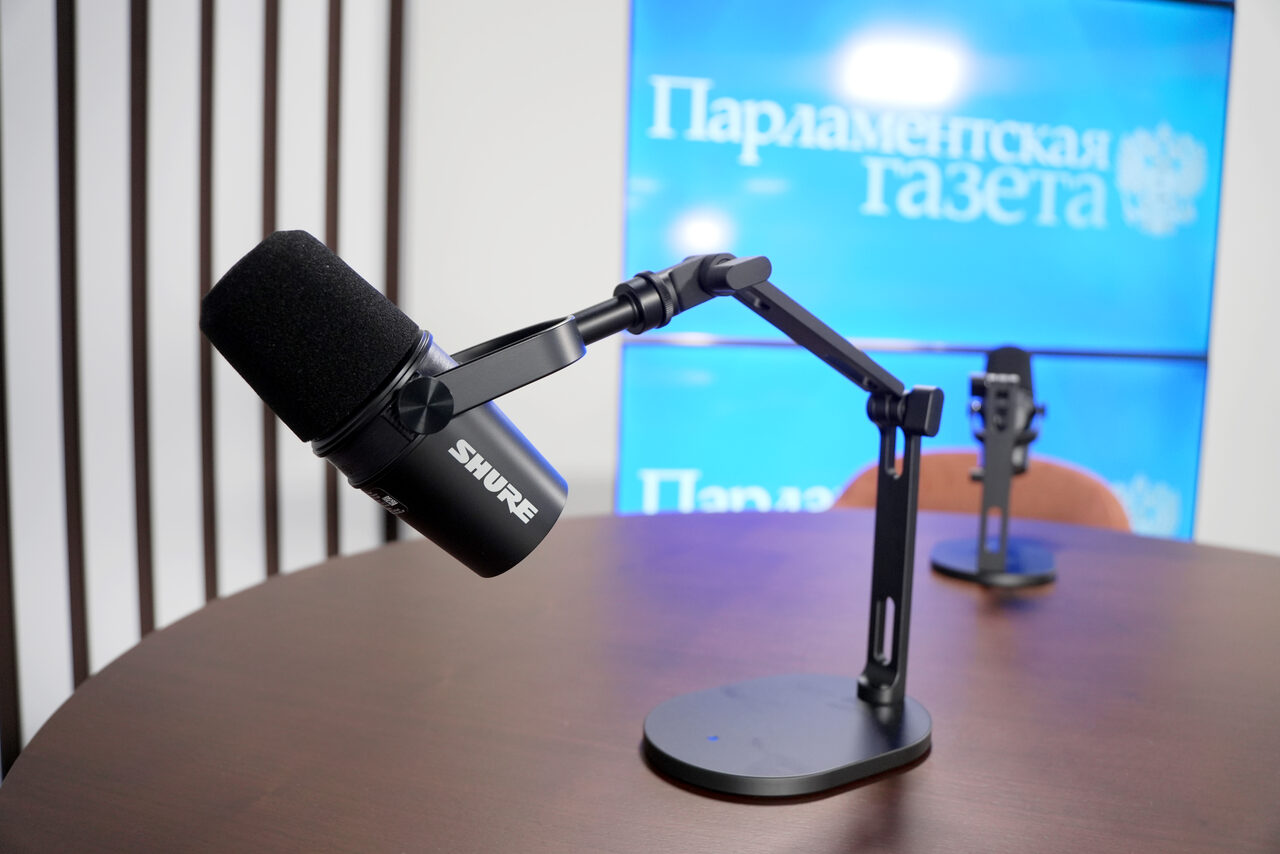 Арендовать Медиацентр Парламентской газеты. Круглый стол с черными микрофонами на стойках, деревянная поверхность. На заднем плане синий экран с текстом "Парламентская газета". Вертикальные черные полосы на стене. Пространство для записи или трансляций.