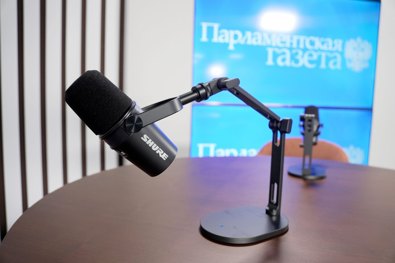 Арендовать Медиацентр Парламентской газеты. Студийное пространство с круглым деревянным столом; на столе установлены микрофоны на настольных стойках; на заднем плане текст "Парламентская газета" на экране; вертикальные планки на стене; современное освещение.