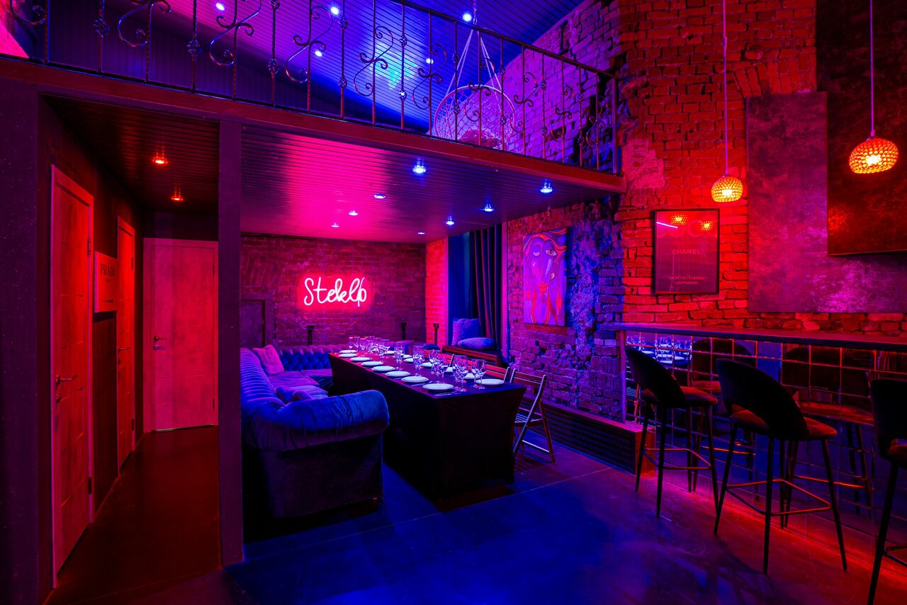 Арендовать МСК Плейс. Лофт Стекло. Loft space with exposed brick walls, featuring a long table set for dining surrounded by chairs and sofas. Ambient red and blue lighting, neon sign on wall, bar area with high stools, and upper mezzanine level visible. Modern industrial style.