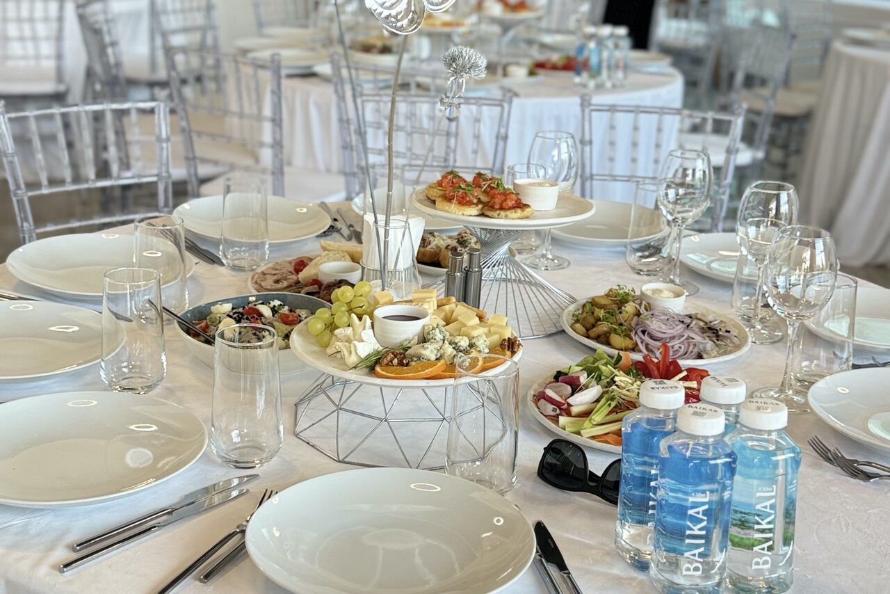 Арендовать Event  Hall ARCOBALENO (Эвент Холл АРКОБАЛЕНО) . Столовая зона с круглым столом, покрытым белой скатертью, сервированная тарелками, приборами и стеклянной посудой. На столе закуски и бутылки воды. Прозрачные стулья, светлый интерьер, круглые столы на заднем плане.