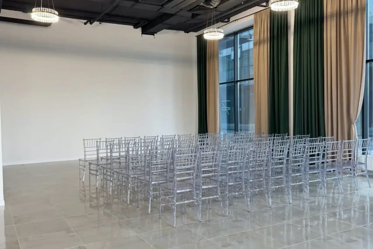 Арендовать Event  Hall ARCOBALENO (Эвент Холл АРКОБАЛЕНО) . Просторное помещение с белыми стенами и большими окнами, закрытыми зелеными и бежевыми шторами. Расставлены прозрачные пластиковые стулья в рядах. Потолочные светильники обеспечивают освещение. Пол выложен светлой плиткой.
