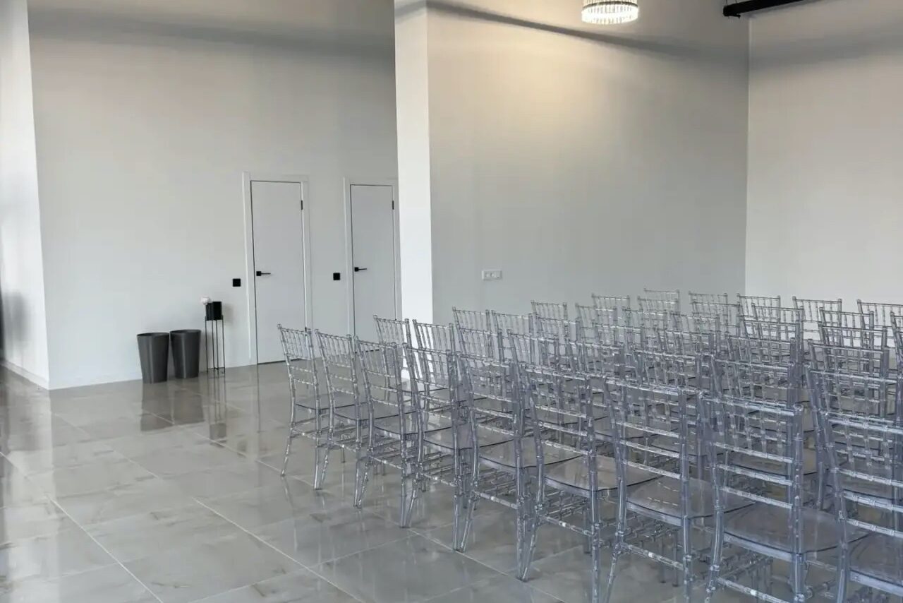 Арендовать Event  Hall ARCOBALENO (Эвент Холл АРКОБАЛЕНО) . Просторная комната с рядами прозрачных стульев, белые стены, светлый плиточный пол, белый потолок с круглыми светильниками. Две двери на стене слева. В углу два мусорных бака.