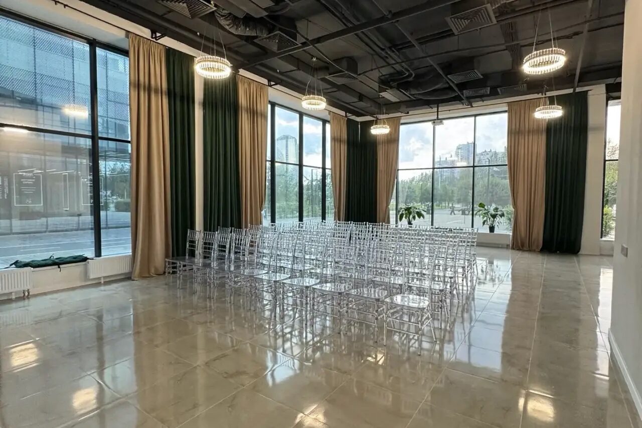Арендовать Event  Hall ARCOBALENO (Эвент Холл АРКОБАЛЕНО) . Просторное помещение с большими окнами и светлыми плиточными полами. Расставлены прозрачные стулья рядами. Потолок открытого типа, висят круглые светильники. На окнах висят длинные шторы зеленого и бежевого цвета. Видны зеленые растения на подоконниках.