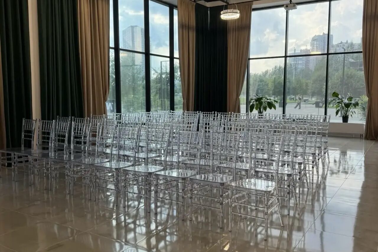 Арендовать Event  Hall ARCOBALENO (Эвент Холл АРКОБАЛЕНО) . Большое помещение с панорамными окнами, несколькими рядами прозрачных стульев, светлый плиточный пол, черные и бежевые шторы, высокий потолок с подвесным светильником, в углу помещения зелёные растения.