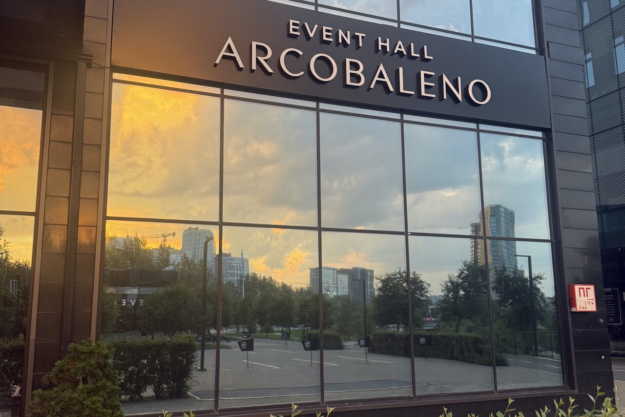 Арендовать Event  Hall ARCOBALENO (Эвент Холл АРКОБАЛЕНО) . Фасад здания с большими зеркальными окнами, отражающими городские пейзажи и закатное небо. Чёрная табличка с текстом "EVENT HALL ARCOBALENO". Состояние фасада ухоженное, внизу зелёные кусты.