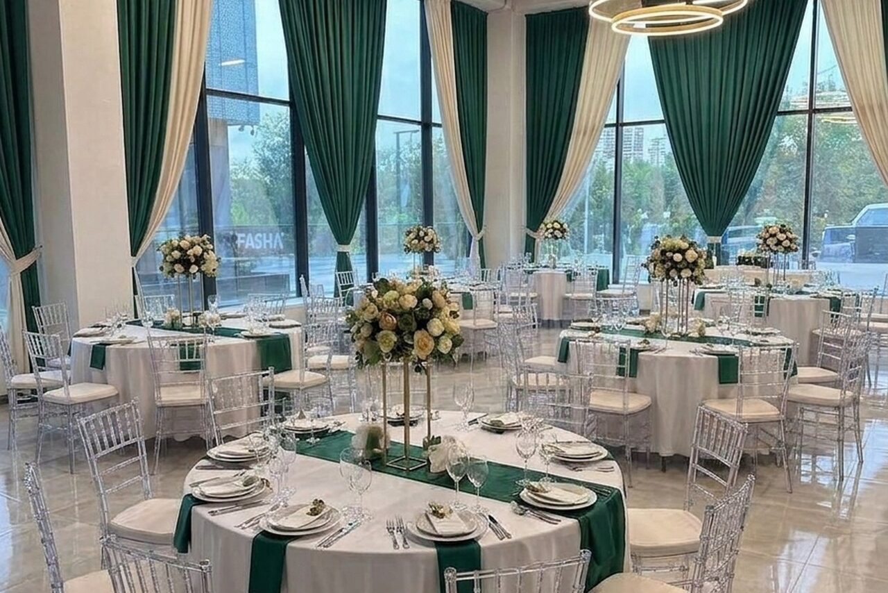 Арендовать Event  Hall ARCOBALENO (Эвент Холл АРКОБАЛЕНО) . Просторный зал с круглыми столами, покрытыми белыми скатертями и зелеными дорожками. Прозрачные стулья вокруг столов. На столах сервировка и цветочные композиции. Высокие окна с зелеными и белыми шторами. Светлые стены и пол, подвесной светильник.