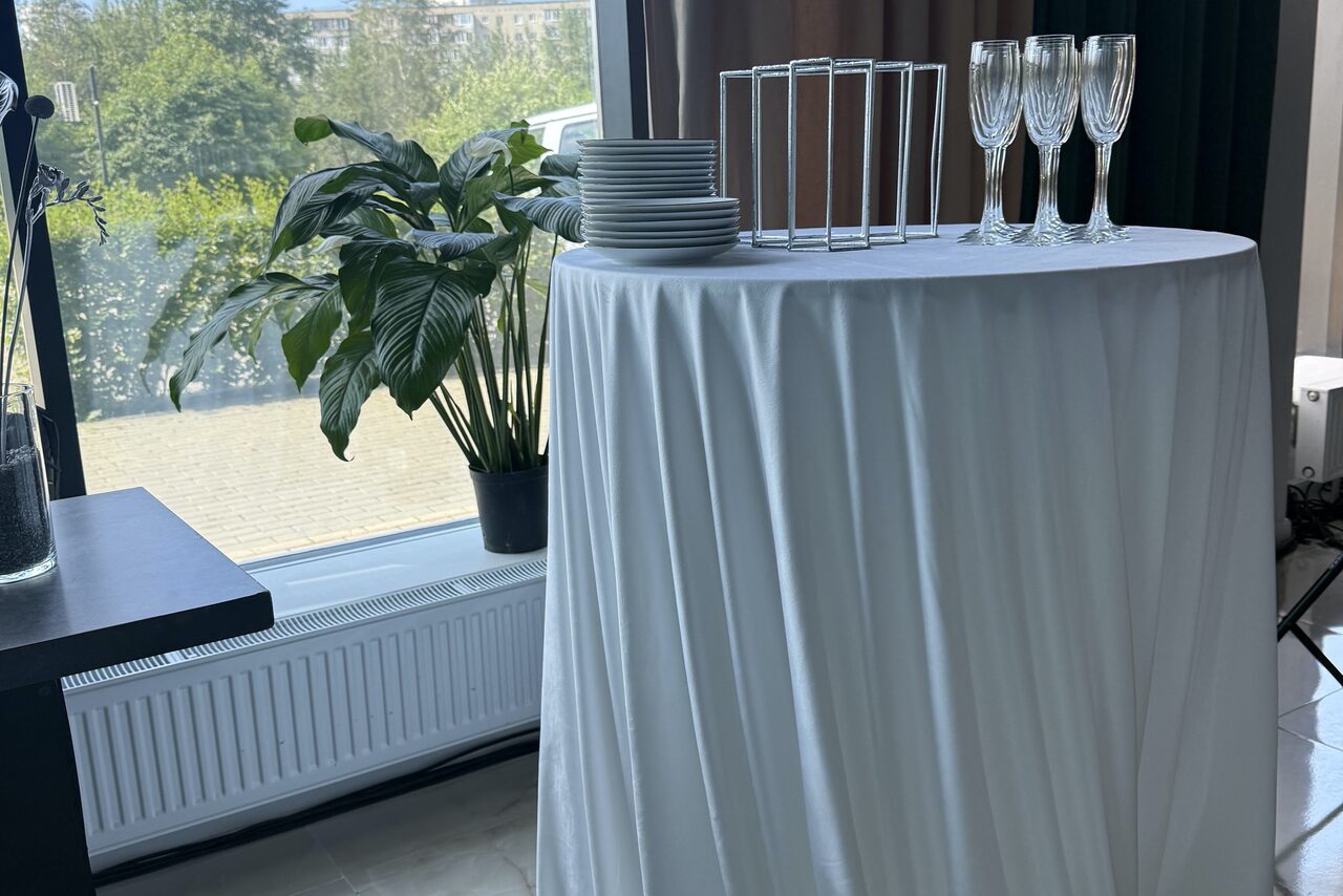 Арендовать Event  Hall ARCOBALENO (Эвент Холл АРКОБАЛЕНО) . Высокий круглый стол, накрытый белой скатертью, с тарелками и бокалами на столешнице. Рядом с окном стоит растение в горшке. Вид из окна на зелень. Светлое помещение с плиточным полом.