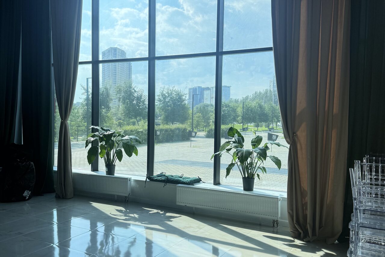 Арендовать Event  Hall ARCOBALENO (Эвент Холл АРКОБАЛЕНО) . Просторное помещение с большими окнами и видом на улицу, занавесками тёмного цвета, плиточным полом и несколькими зелеными растениями в горшках на подоконнике. На полу видны прозрачные стулья и радиаторы отопления под окнами.