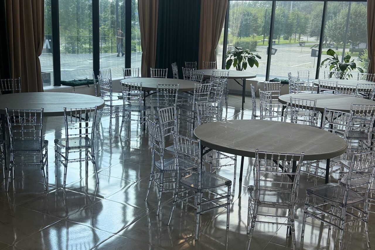 Арендовать Event  Hall ARCOBALENO (Эвент Холл АРКОБАЛЕНО) . Просторное помещение с большими окнами, круглыми столами и прозрачными стульями. Полы плиточные, стены с тяжелыми шторами. Растения на подоконниках. Естественное освещение от панорамных окон.