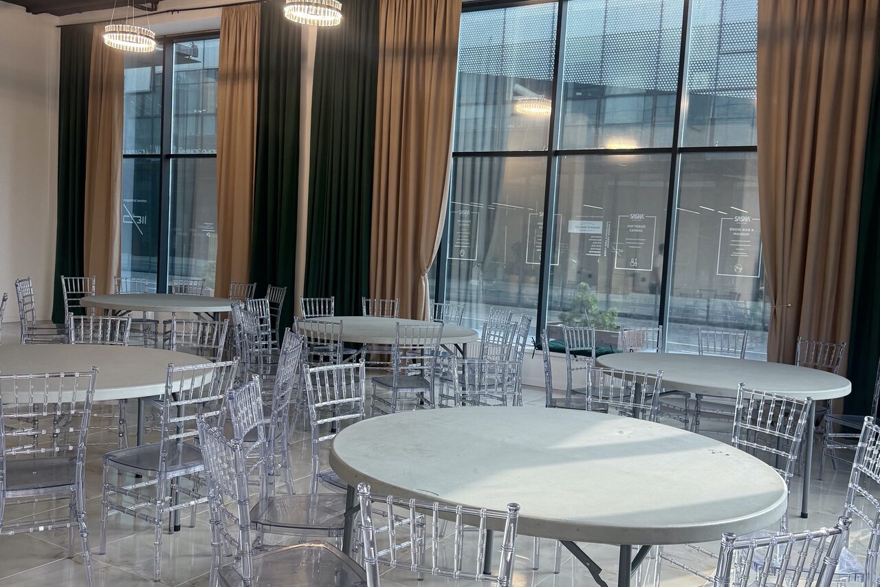 Арендовать Event  Hall ARCOBALENO (Эвент Холл АРКОБАЛЕНО) . Просторное помещение с круглыми столами и прозрачными стульями. Большие окна со шторами, через которые видно здание напротив. Потолочные светильники круглой формы. Пол светлый, стены нейтральные.
