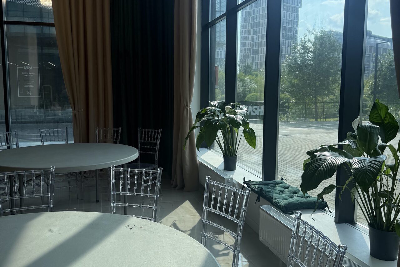 Арендовать Event  Hall ARCOBALENO (Эвент Холл АРКОБАЛЕНО) . Просторная комната с большими окнами, круглые столы и прозрачные стулья, светло-зелёные и тёмные шторы, на подоконнике подушки и зелёные растения в горшках.