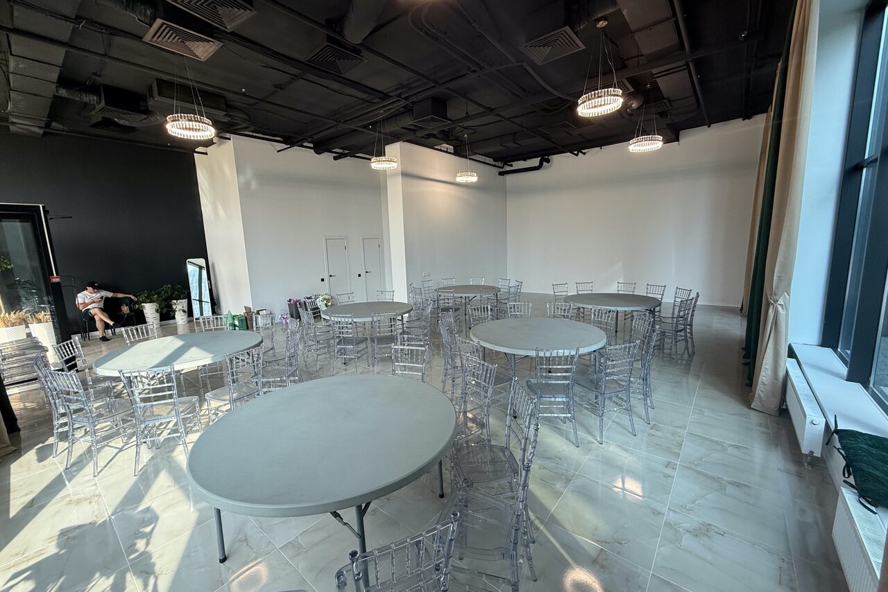 Арендовать Event  Hall ARCOBALENO (Эвент Холл АРКОБАЛЕНО) . Просторное помещение с белыми стенами и светлым плиточным полом. Расставлены круглые столы и прозрачные стулья. Потолок черный, с подвесными светильниками. Большие окна слева, двери и зеркала на заднем плане, растения в уголке.