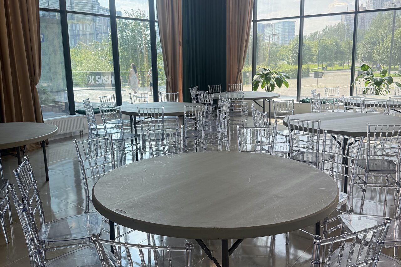Арендовать Event  Hall ARCOBALENO (Эвент Холл АРКОБАЛЕНО) . Просторное помещение с круглыми столами и прозрачными стульями, большие окна до пола с видом на улицу, светлые полы, коричневые и тёмно-зелёные шторы, растения у окон, яркое естественное освещение.