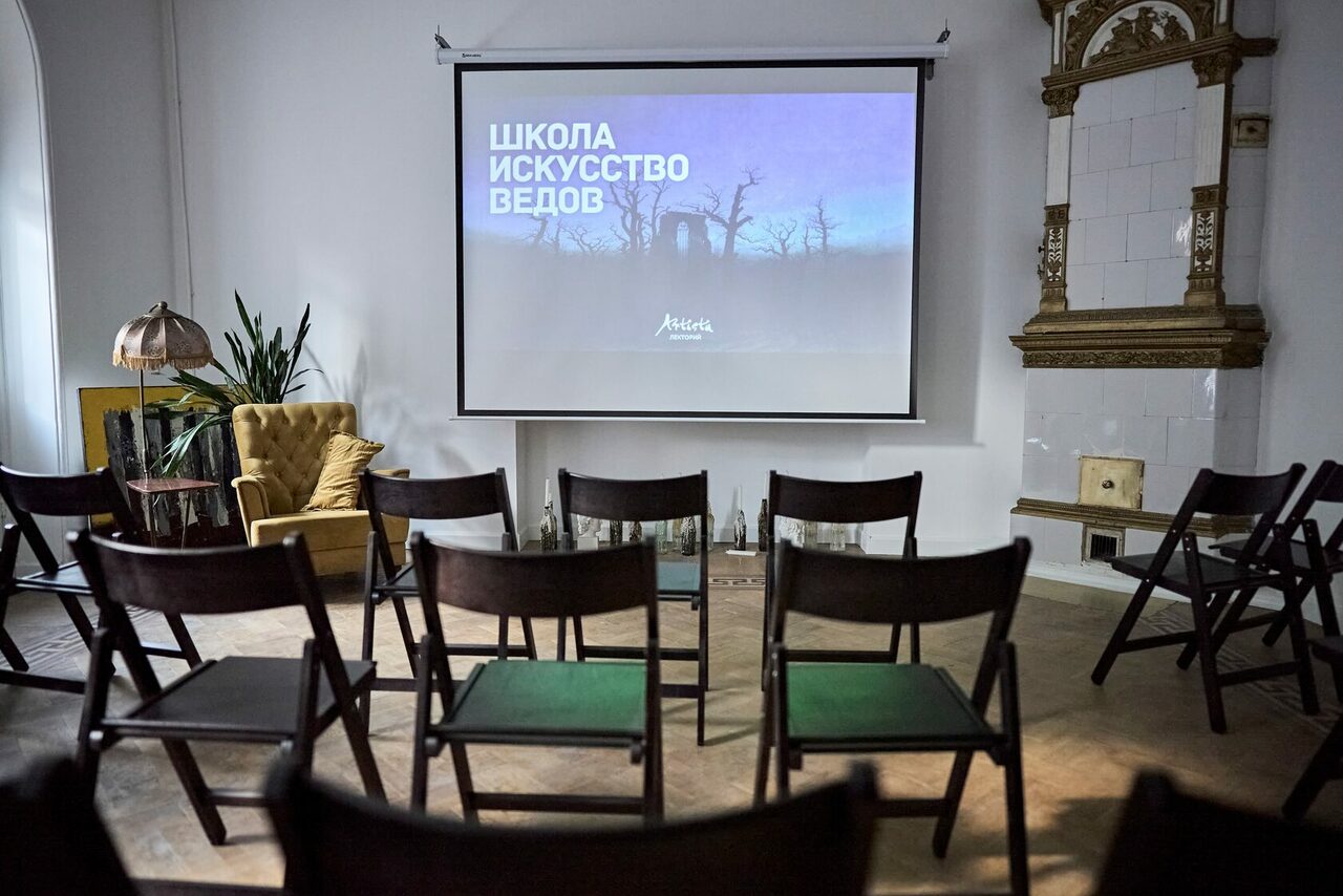 Арендовать Артиста Лекторий. Пространство с деревянным полом, белыми стенами и экраном для проектора. Ряды черных складных стульев, на экране текст. У стены кресло, зелёное растение и светильник. Справа здание с декоративной плиткой.