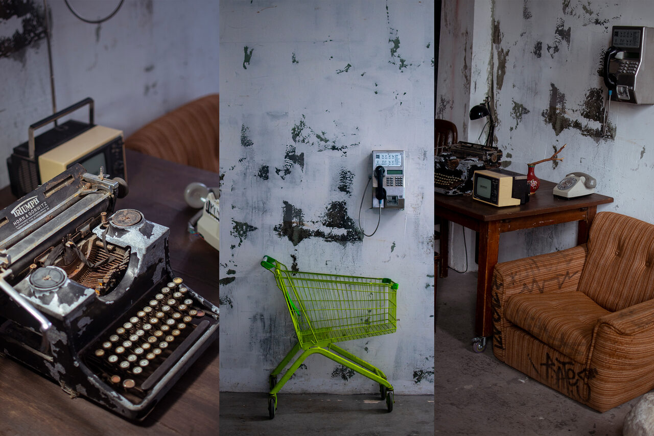Арендовать Фотостудия Грани. Пространство с индустриальной обстановкой: письменный стол с пишущей машинкой и старыми телефонами, рядом кожаное кресло. На стене потертая краска, висят телефон-автомат и счётчик. В углу тележка для покупок. Цвета тёмные, материалы дерева и металла.
