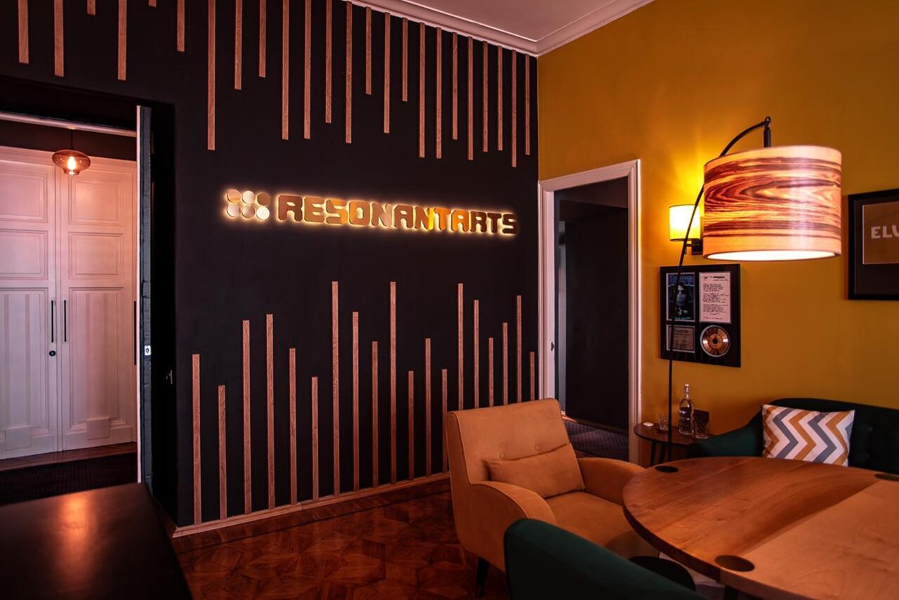 Арендовать Resonant Arts – студия звукозаписи и видео съёмки. Современное пространство с черной стеной и деревянными вставками, надписью "Resonamate". Оранжевое кресло и деревянный стол. Желто-оранжевая стена с постером и квадратным светильником. Паркетный пол и круглые светильники.