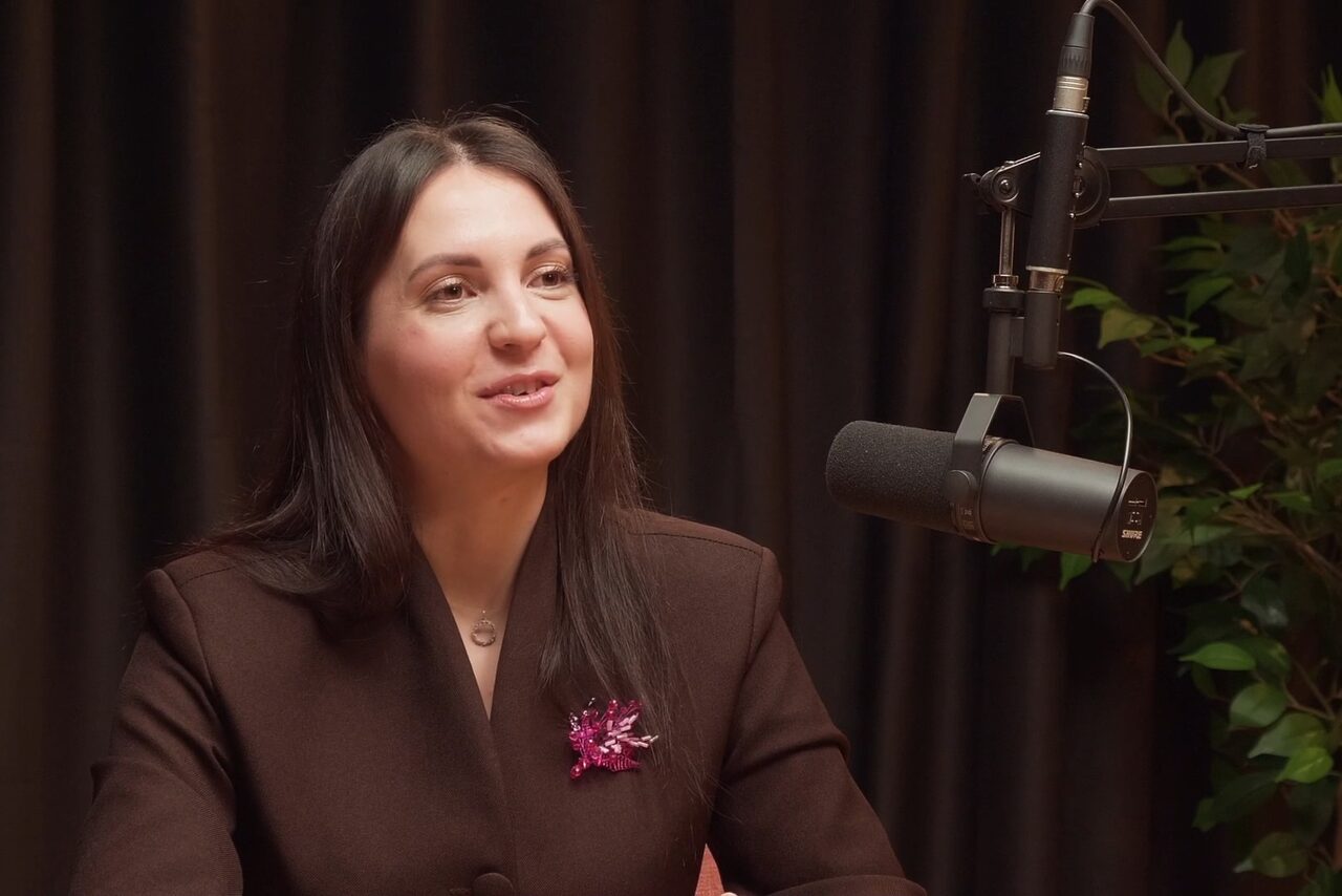 Арендовать Подкастная А40. Студийное пространство с затемненной драпировкой, крупная микрофонная установка, на заднем плане растение. Женщина в темном пиджаке с розовой брошью сидит перед микрофоном. Освещение мягкое.
