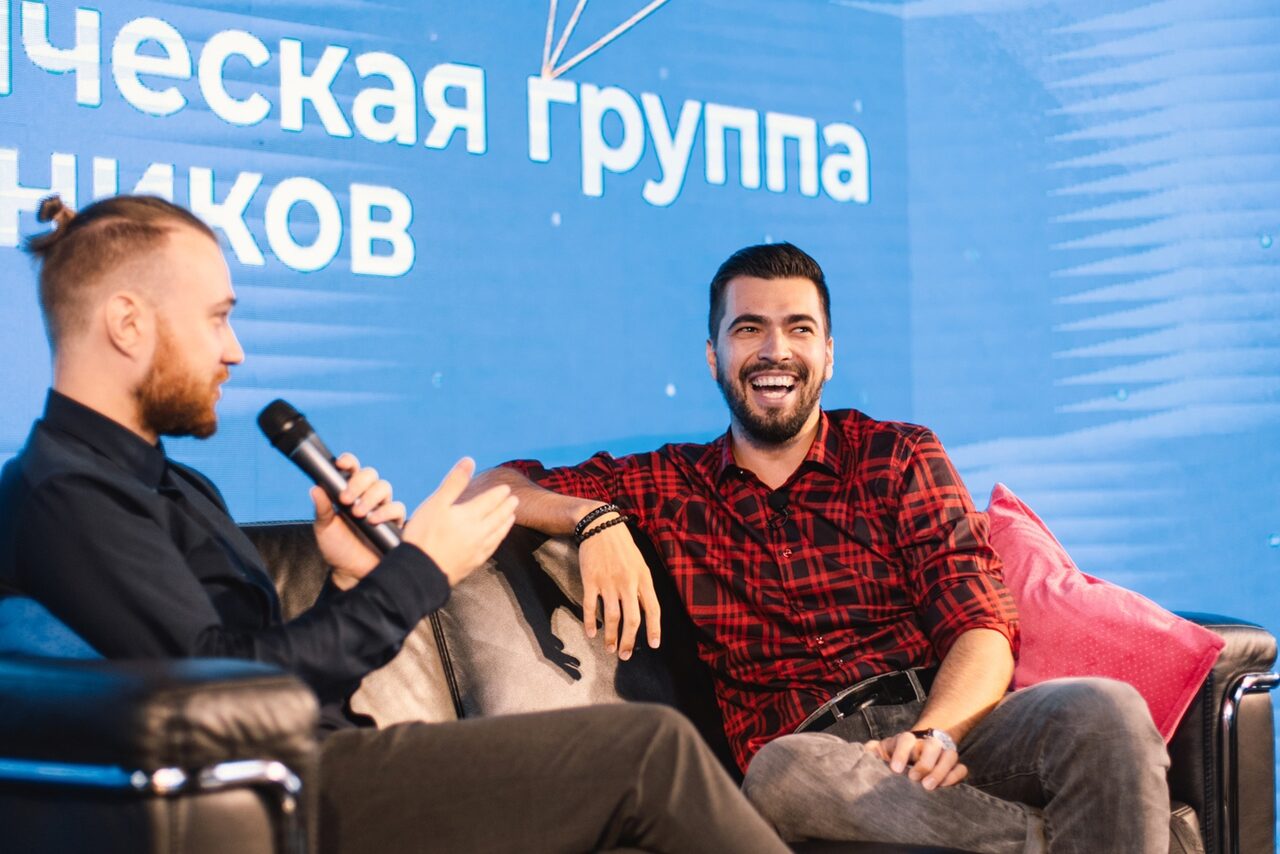 Арендовать Конференц-зал площадью 313 м² до 150 гостей. Гостиная зона с двумя мужчинами на кожаном диване. Мужчина справа в красно-черной рубашке, мужчина слева с микрофоном. На фоне синий экран с текстом на русском. Подушки на диване.