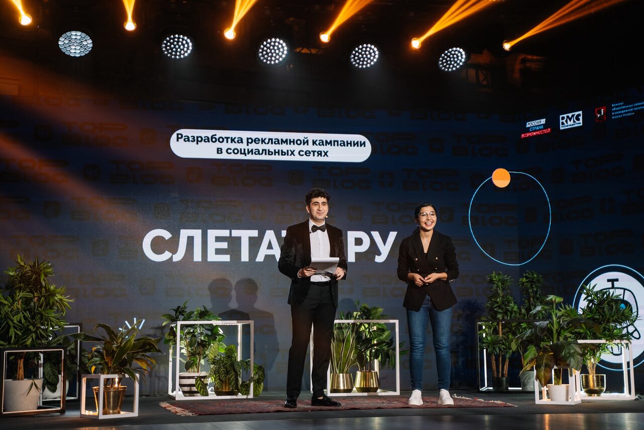 Арендовать Конференц-зал площадью 313 м² до 150 гостей. Сцена с двумя людьми, фоном используется большой экран с надписью "Разработка рекламной кампании в социальных сетях". На сцене много зелёных растений в горшках. Освещение тёплое, сверху направлены светодиодные лампы.
