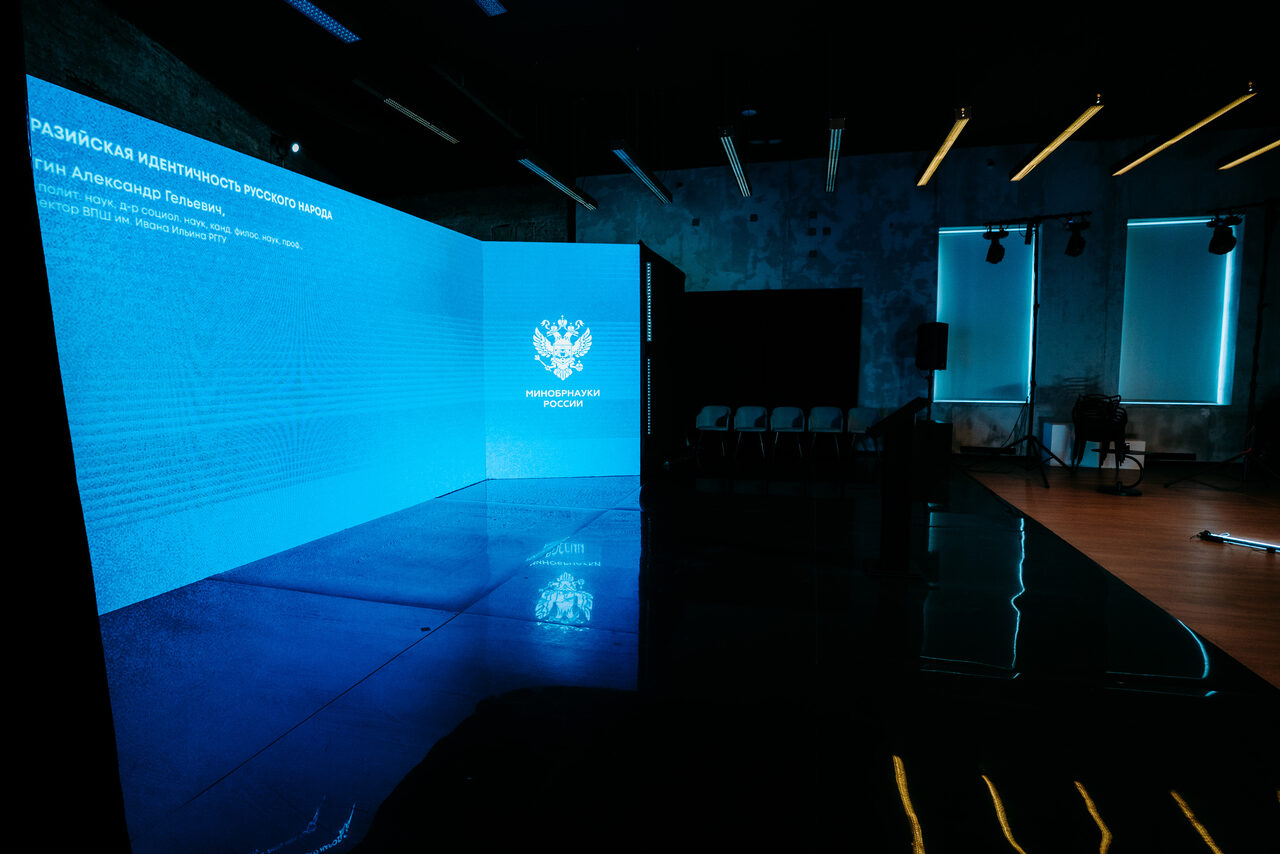 Арендовать Конференц-зал площадью 313 м² до 150 гостей. Современное просторное помещение с темными стенами и потолком. Большой экран с голубым цифровым отображением и текстом. Ряды черных стульев, деревянный пол. Яркое потолочное освещение и светодиодная подсветка.