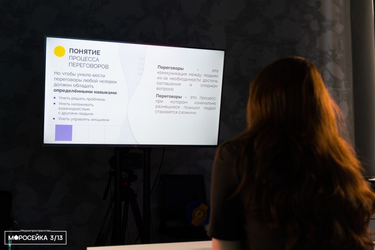 Арендовать Конференц-зал площадью 313 м² до 150 гостей. Пространство для встреч с затемненной обстановкой, столом и стулом. На стене установлен экран на штативе с текстом о процессе переговоров. Видна тёмная стена и сидящая человек спиной. Освещение мягкое, приглушенное.