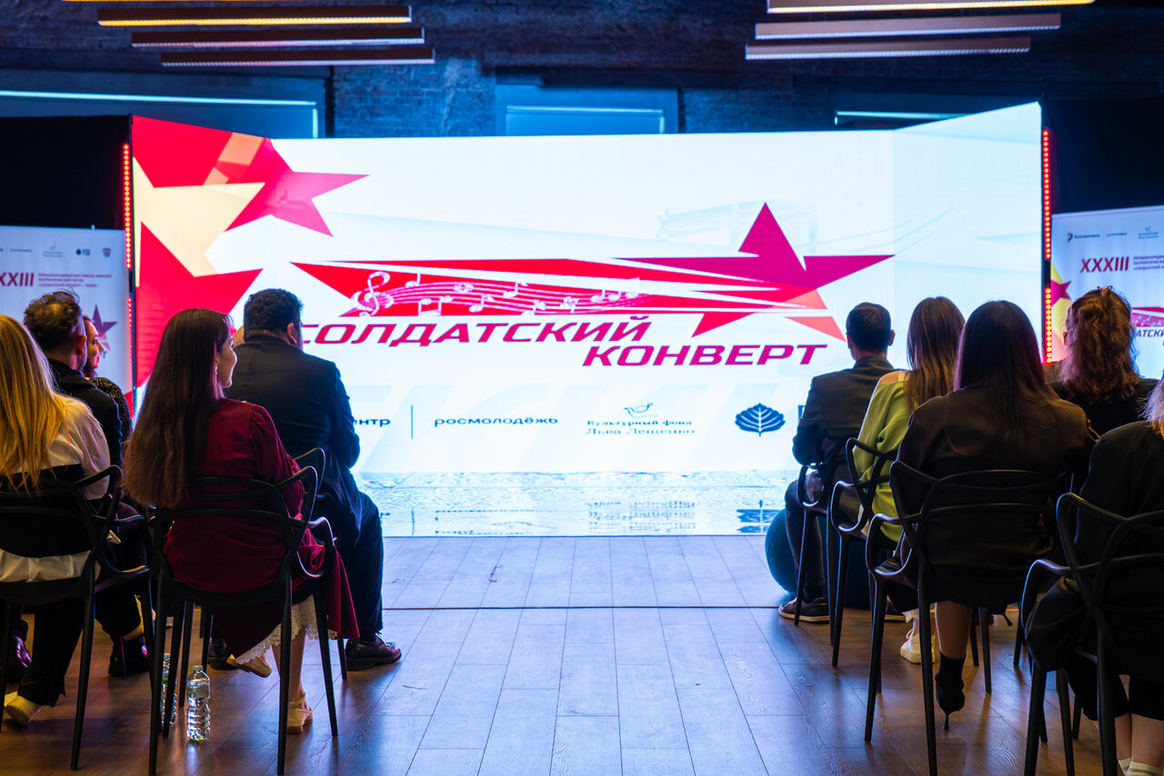 Арендовать Конференц-зал площадью 313 м² до 150 гостей. Две группы людей сидят на стульях перед большим экраном с красно-белой графикой и текстом «Солдатский конверт». Стены из темного кирпича, потолочное освещение линейного типа. Пол деревянный, присутствуют темные элементы интерьера.