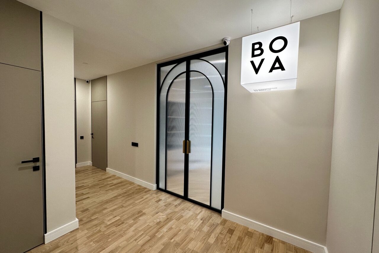 Арендовать Лекторий на 53 этаже Москва-Сити - 100 м², до 35 человек. Коридор с деревянным полом и светлыми стенами, стеклянная дверь с арочной конструкцией и черной рамой, рядом светящийся белый куб с черной надписью "BOVA". На дверях черные ручки. Освещение встроенное.