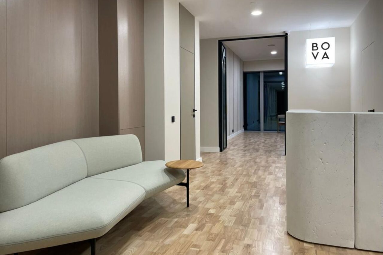 Арендовать Лекторий на 53 этаже Москва-Сити - 100 м², до 35 человек. Современное офисное пространство с паркетным полом, светлыми стенами и потолком. Слева мягкий диван с небольшим встроенным столиком, справа бетонная стойка. В глубине виден стеклянный вход. Освещение точечное. На стене знак с текстом "BOVA".